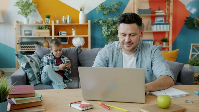 Modern home office setup with father on laptop and child using a smartphone on the sofa. Werk-privébalans: roosters, verlof en grenzen stellen